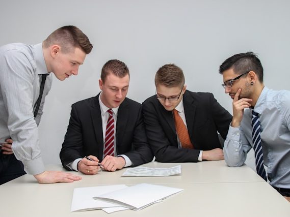 Vier Männer in Anzügen diskutieren über Dokumente an einem Tisch.