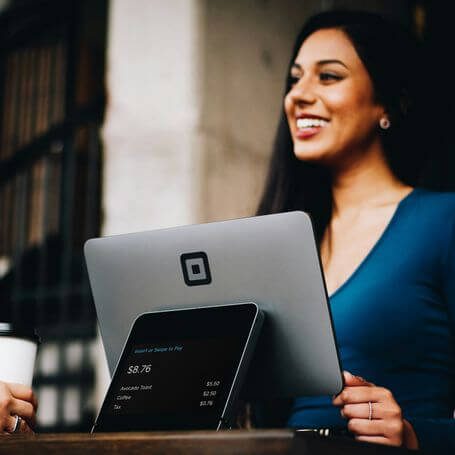 Eine Frau in blauer Bluse lächelt, während sie ein Tablet und eine Tasse Kaffee hält.